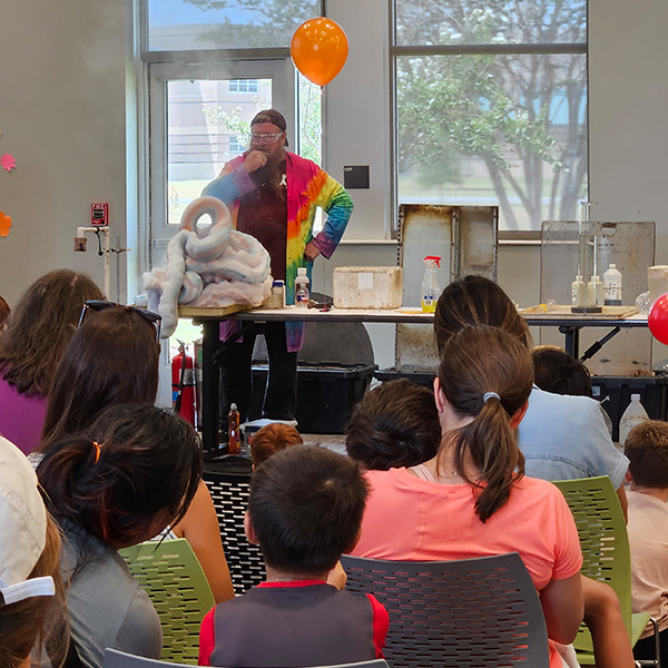 Chemistry Roadshow host presenting a table full of science demonstrations to a group of children. 